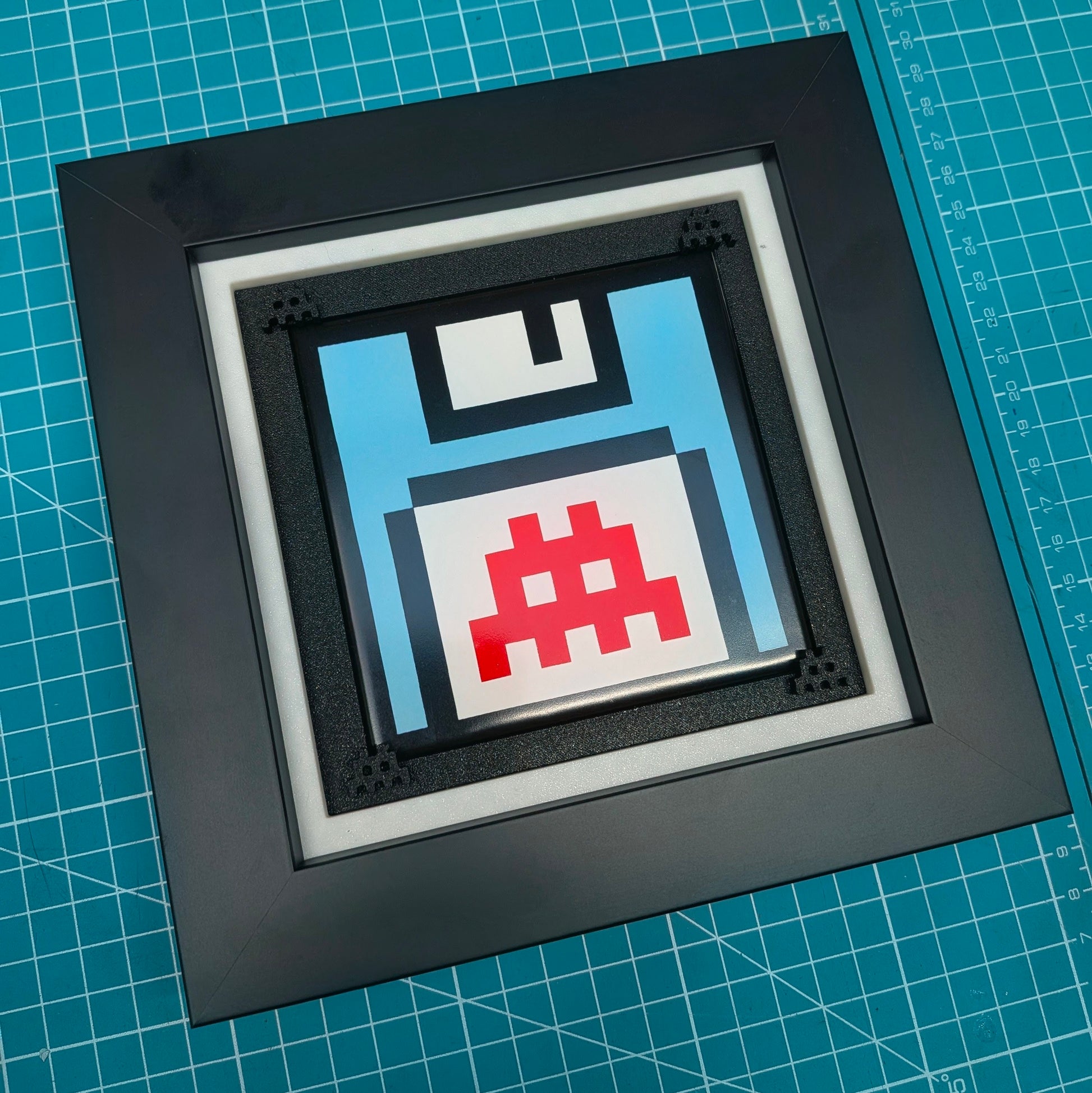 A square black shadowbox frame containing a pixel-art floppy disk design in light blue, white, and red, displayed on a green cutting mat.