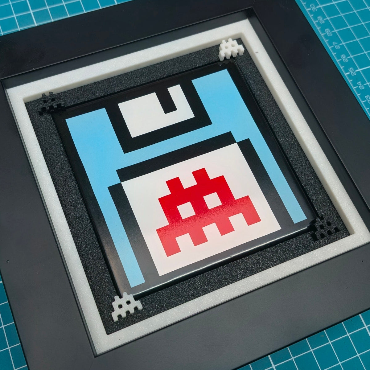 A square framed art print featuring a pixelated retro floppy disk design in light blue, black, white, and red, displayed on a blue gridded cutting mat.