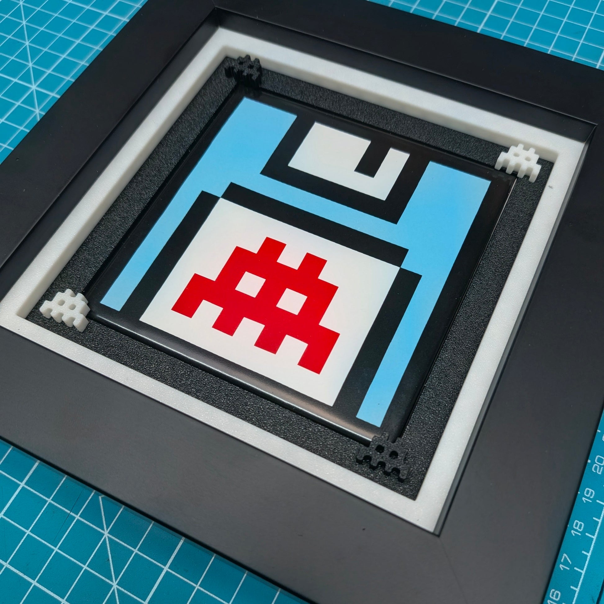 A square framed pixel art print depicting a retro floppy disk with a red hashtag symbol on a light blue and white background, set inside a black frame with white matting, placed on a blue cutting mat.