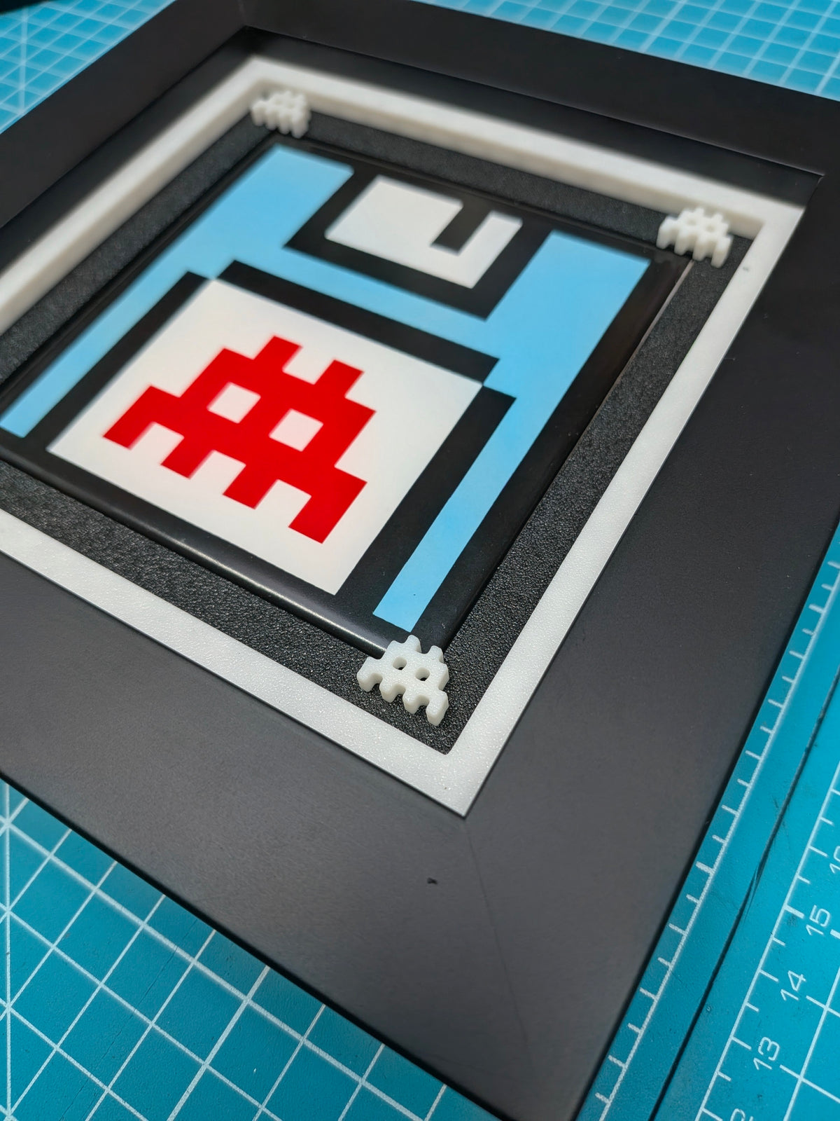 A square framed pixel art print depicting a retro floppy disk with a red hashtag symbol, displayed on a blue gridded cutting mat.