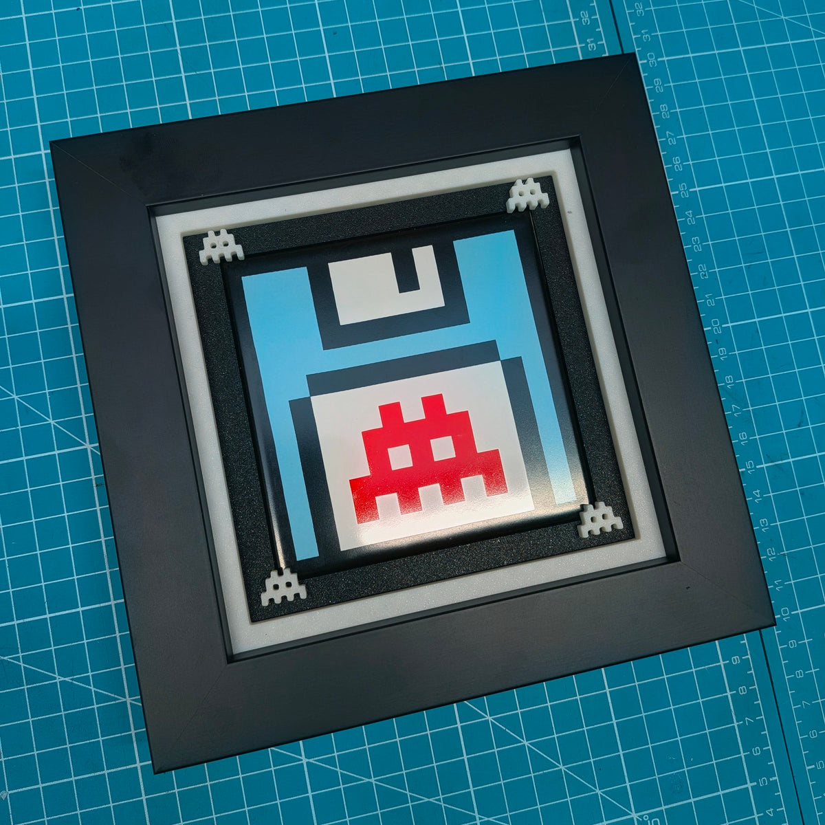 A square black shadowbox frame containing a pixel-art retro floppy disk design in light blue, white, and red, displayed on a blue cutting mat.