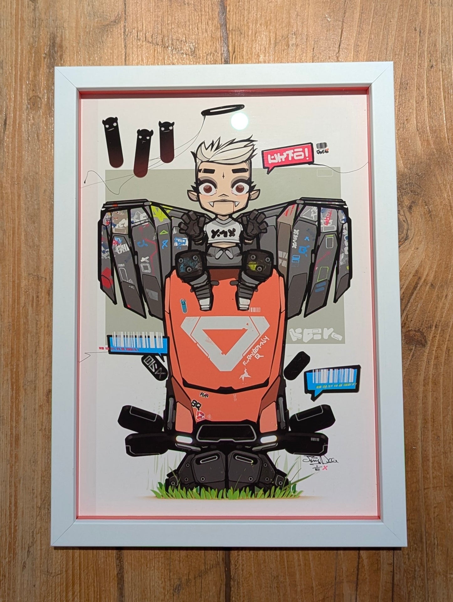 frame anime style character sat on red robot, the character has grey wings and vampire teeth, the frame is white with a colour matching slip. picture is placed on wooden table
