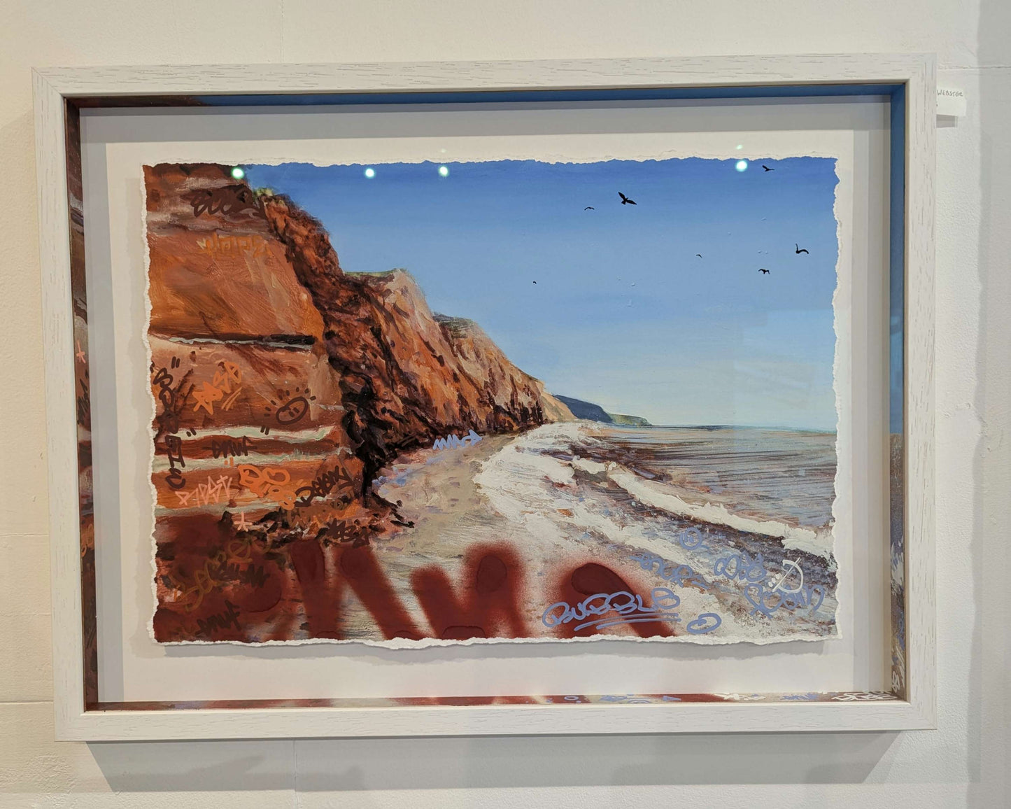 white frame against white wall, artwrok is by joe webster, its shows red cliffs down by the sea, graffiti tags are drawn on the cliffs but in matching colours