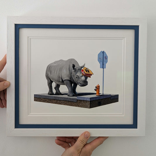 Framed artwork of a rhinoceros with a small figure and blue box on a white background being held.