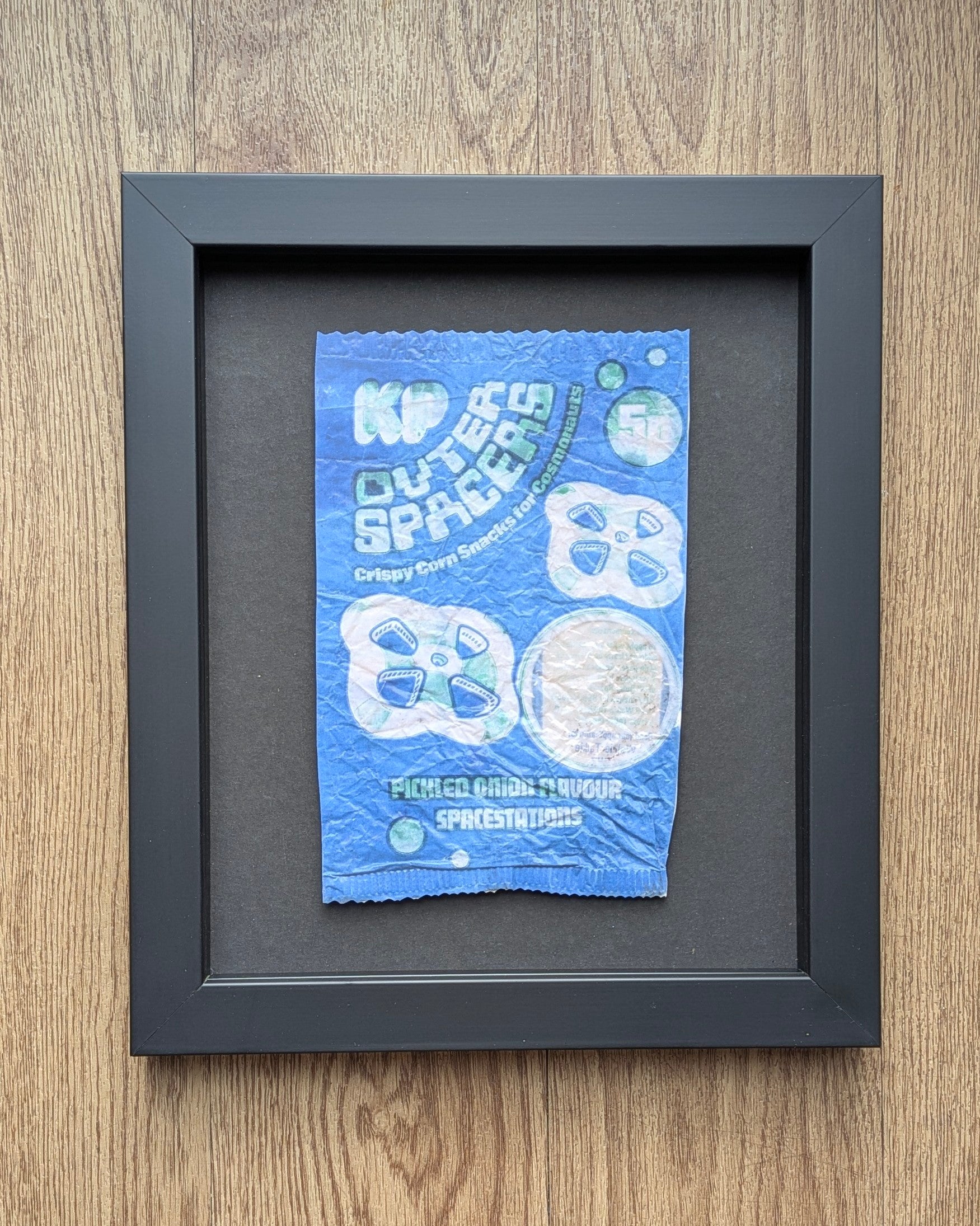 Framed blue and white kp outer spacers crisp packet packaging on a wooden surface