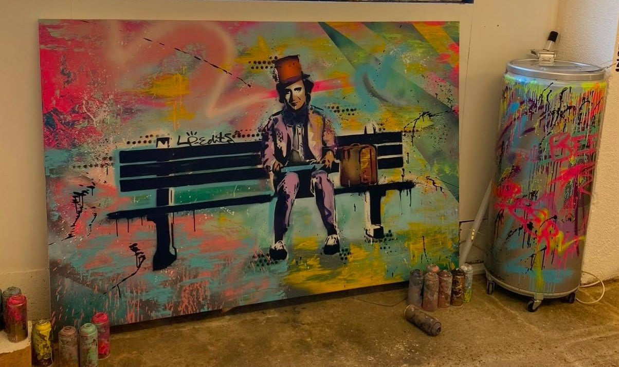 A large canvas painting featuring a seated man on a bench with a colorful graffiti-style background, displayed indoors next to a matching spray-painted cylindrical container.
