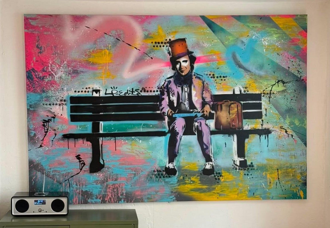 A large colorful wall art painting hangs on a beige wall above a small gray speaker. The artwork depicts a seated figure on a bench with a suitcase, rendered in expressive graffiti-style brushstrokes of pink, blue, yellow, teal, black, and white.