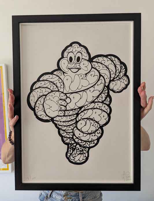 Framed illustration of the cartoon michelin man, black balloon style character with pure drawn all over the body, black pen on white paper held by two hands.