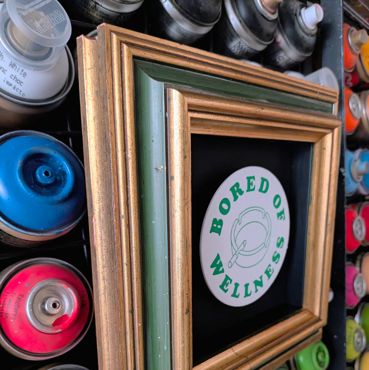 A square framed wall art print featuring a white circular sticker design with green text reading 'BORED OF WELLNESS' and a simple line drawing, mounted inside a layered wooden frame with gold, green, and brown tones. Spray paint cans are visible in the background.