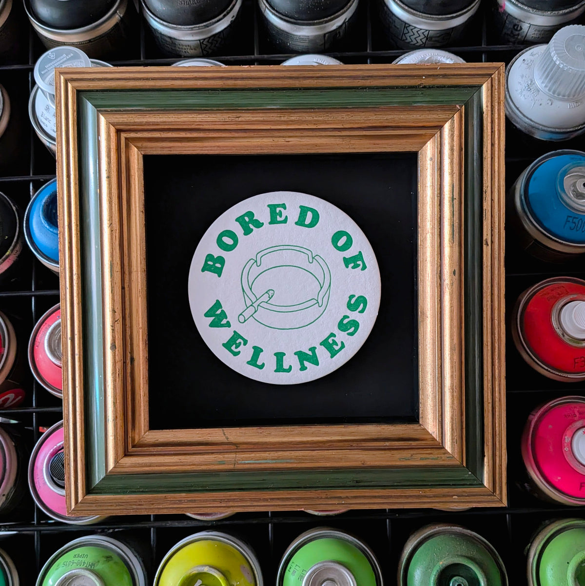 A square framed sticker or art print featuring a white circular design with green text reading 'BORED OF WELLNESS' and a simple green illustration, set in a rustic multicolor wooden frame. The frame is displayed against a background of colorful spray paint cans.