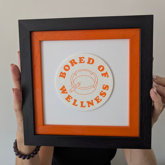 Framed artwork bored of wellness beer mat, green text with image of ashtray and lit cigarette, beer mat is float mounted on a double frame, internal frame is orange and black frame, held by a person