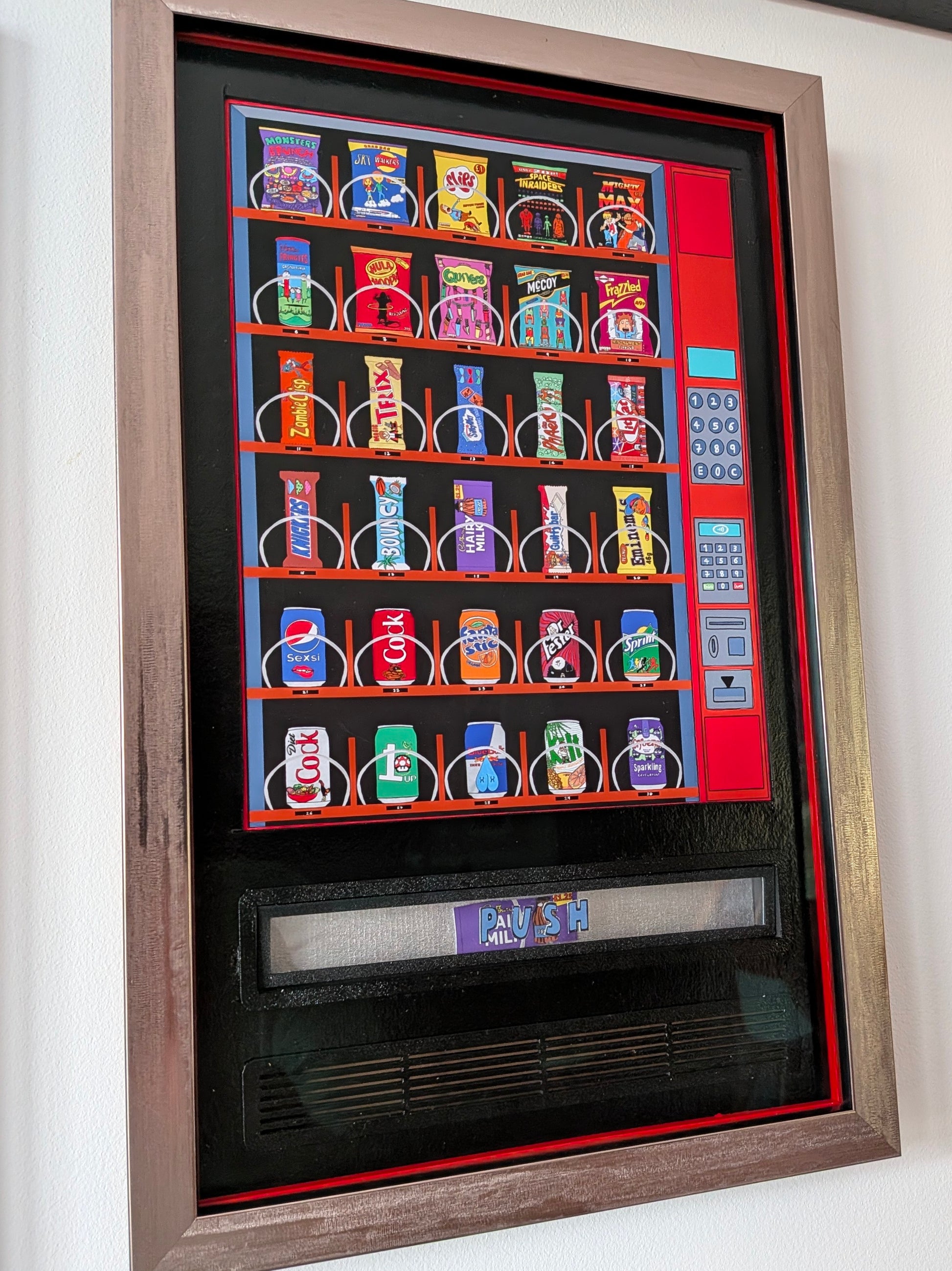 A framed pop-art style print depicting a retro vending machine with colorful snack and drink cans, mounted on a light-colored wall.