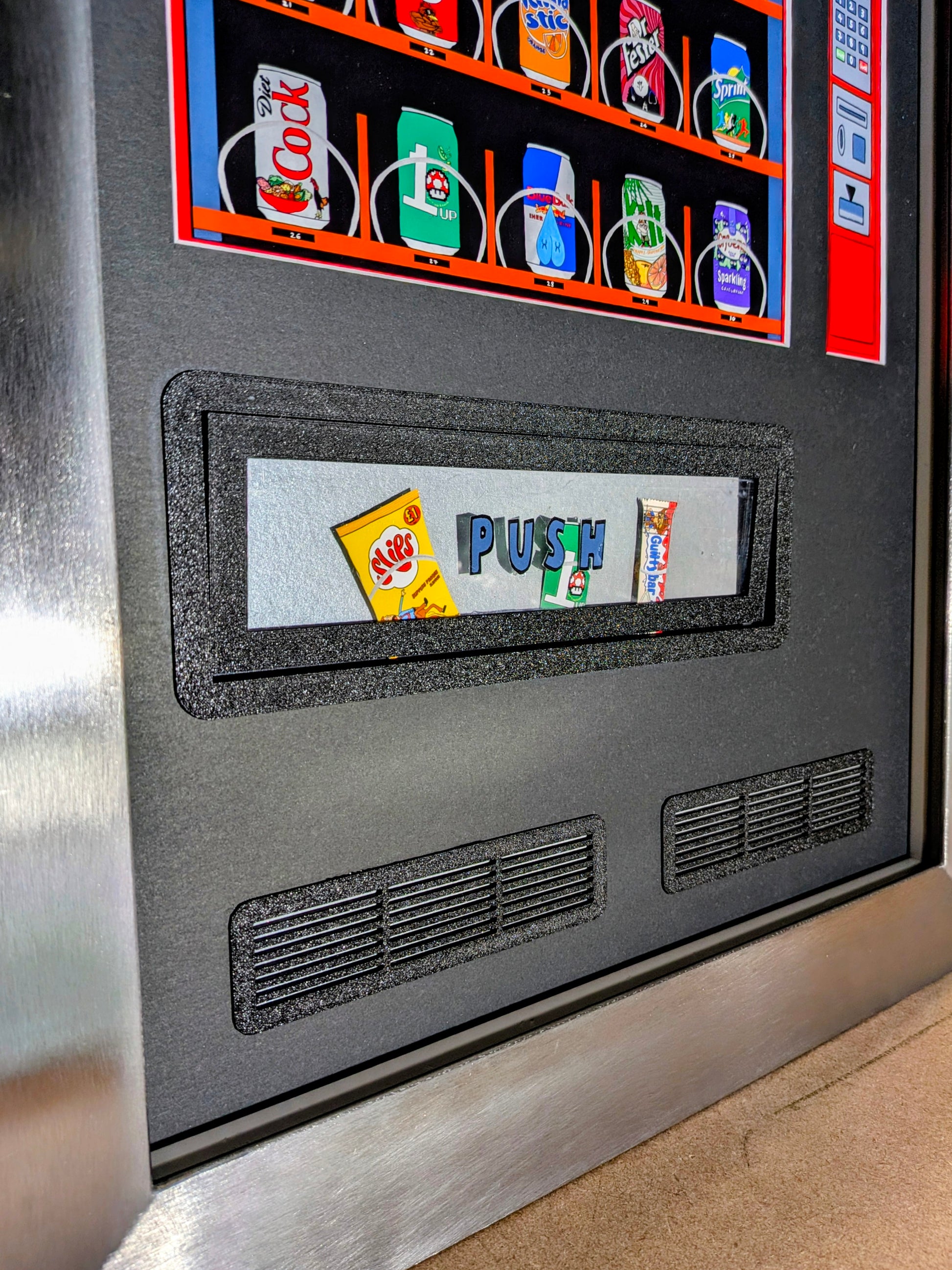 Close-up of a vending machine display with rows of assorted canned drinks and snacks, plus a central coin slot area with the word PUSH and small snack illustrations. The machine has a dark gray front panel with metallic silver trim and ventilation grilles.