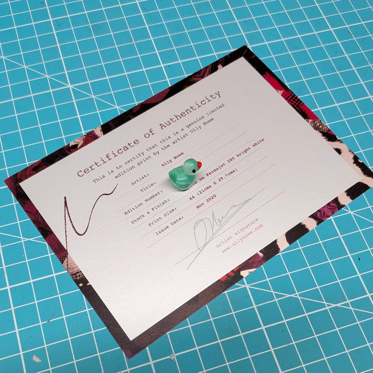 A Certificate of Authenticity card with a patterned border, placed on a blue grid cutting mat. The card features a small green rubber duck in the centre and printed text with placeholder details.