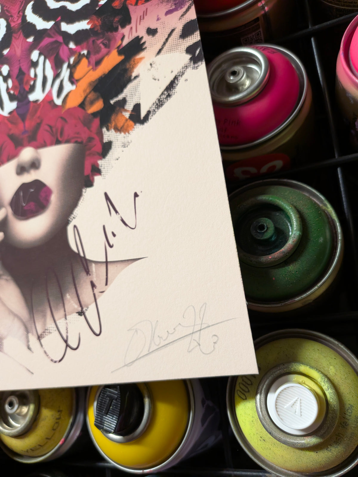 A close-up of a printed card or art print featuring a stylized portrait with bold red, black, orange, and white brushstrokes, overlaid with handwritten signatures. Spray paint cans in pink, green, yellow, and black are visible around it.
