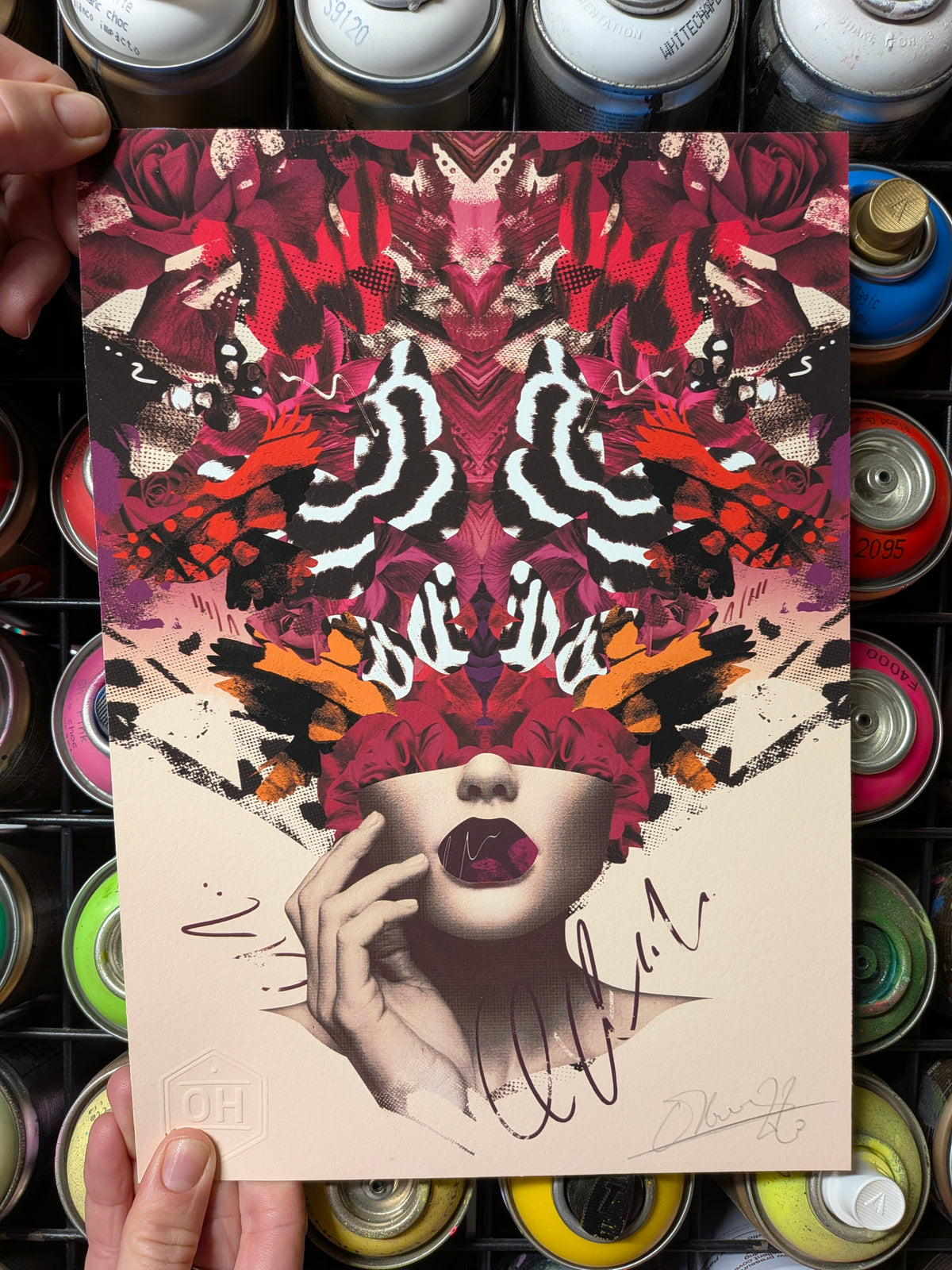 A hand holds a square art print featuring a symmetrical, kaleidoscopic portrait of a woman's face with red flowers and abstract brushstrokes. The background shows a rack of colorful spray paint cans.