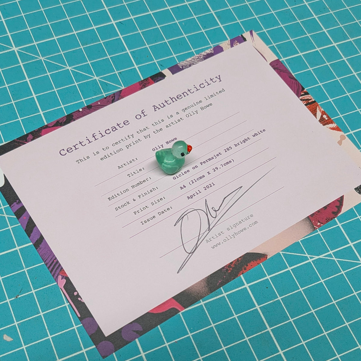 A white Certificate of Authenticity document with printed text and a small green teardrop-shaped charm resting on it, placed on top of a colorful patterned sheet over a turquoise cutting mat background.