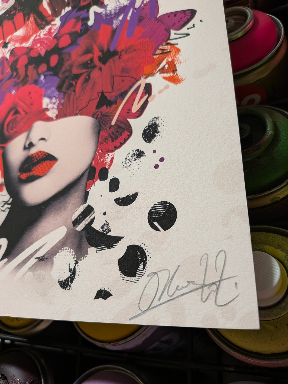 A close-up of a printed artwork featuring a stylized woman's face with bold red, purple, orange, black, and white abstract brushstrokes and paint splatters. The print is photographed over a stack of colorful spray paint cans.