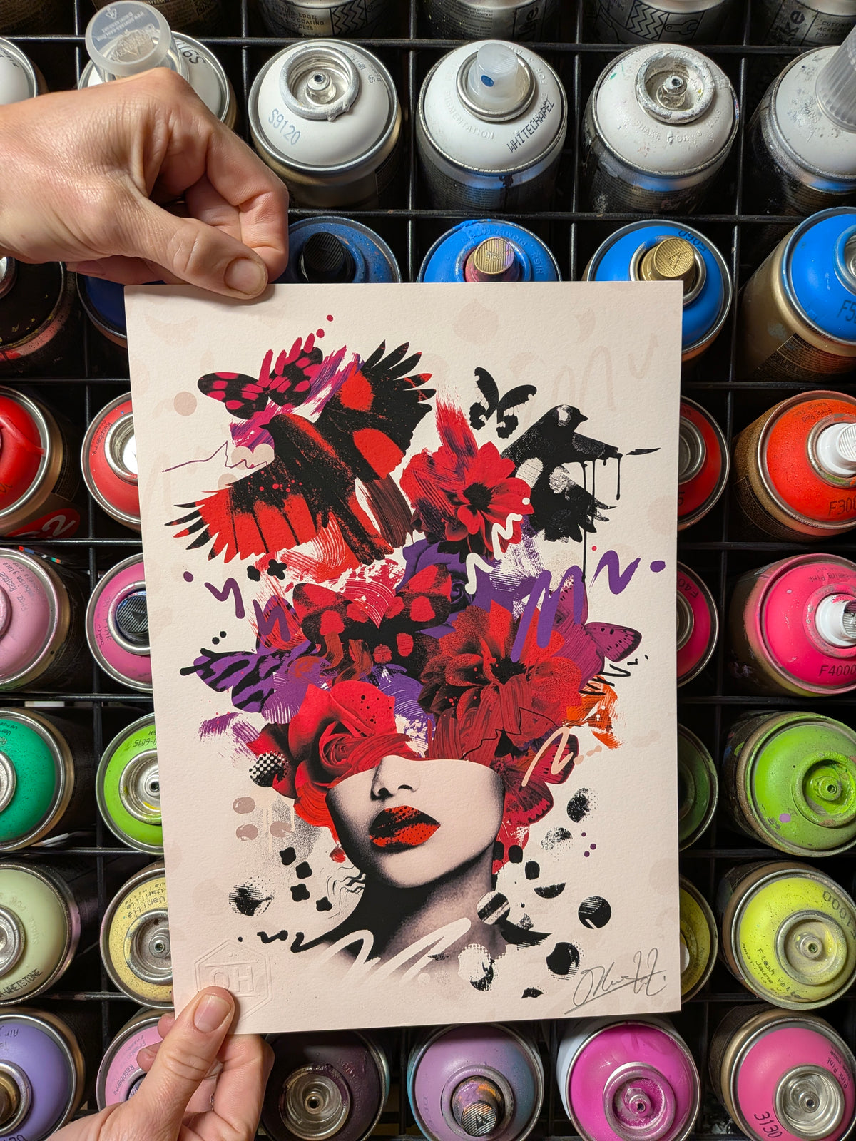A hand holds a square art print featuring a stylized portrait of a woman with red lips and a large floral, bird, and paint-splatter headpiece in red, purple, black, and white. The print is photographed in front of a grid of colorful spray paint cans.
