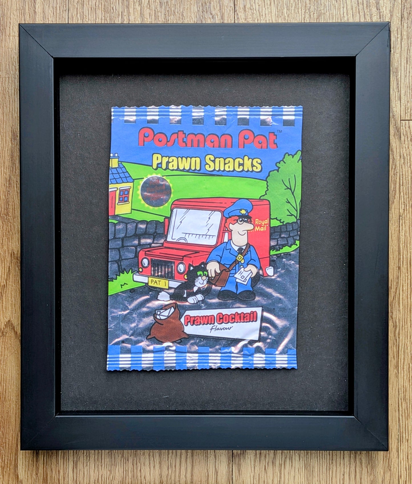 Framed illustration of a cartoon book titled 'Postman Pat Prawn Snacks' on a wooden surface.