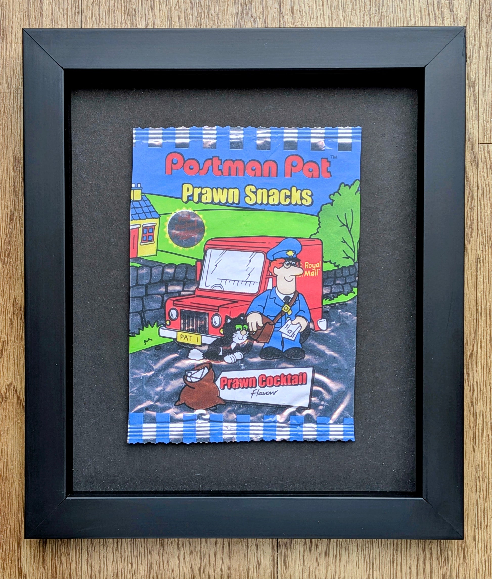 Framed illustration of a cartoon book titled 'Postman Pat Prawn Snacks' on a wooden surface.