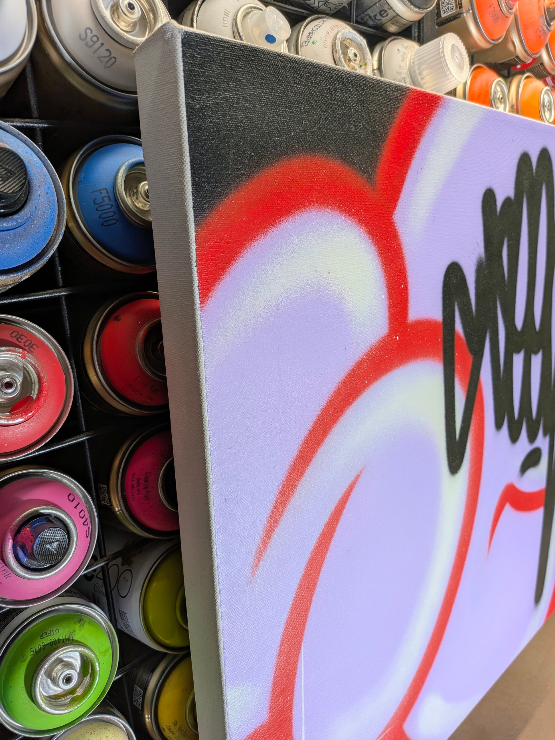 A square canvas print featuring graffiti-style artwork with red, white, black, and purple paint, shown leaning against a rack of colorful spray paint cans.