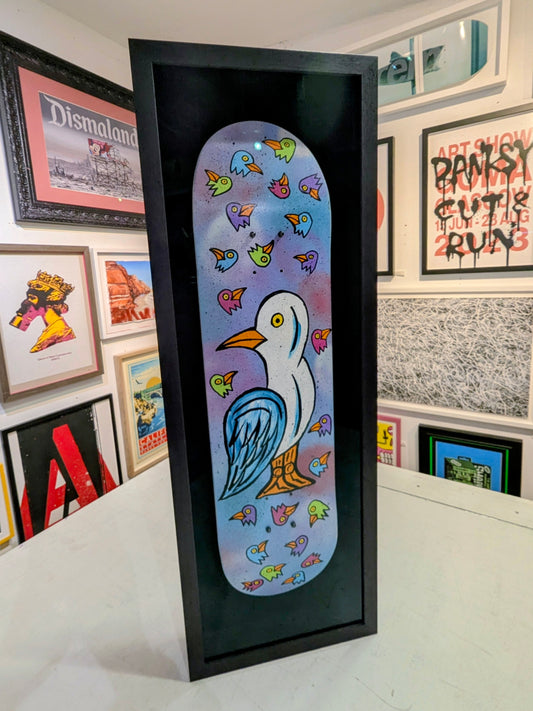 Framed skateboard  with artwork of a colourful bird with bird head designs on a blue background, displayed among other framed pictures.