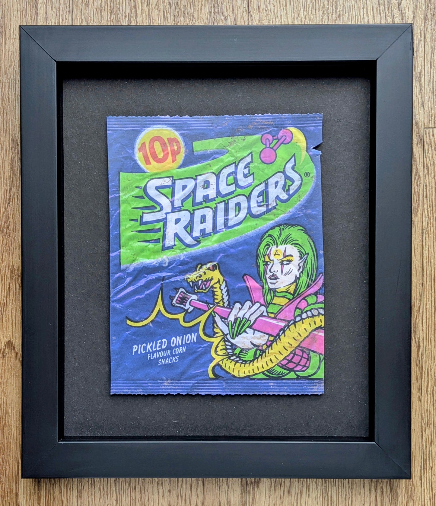 Framed vintage Space Raiders candy packaging on a wooden surface