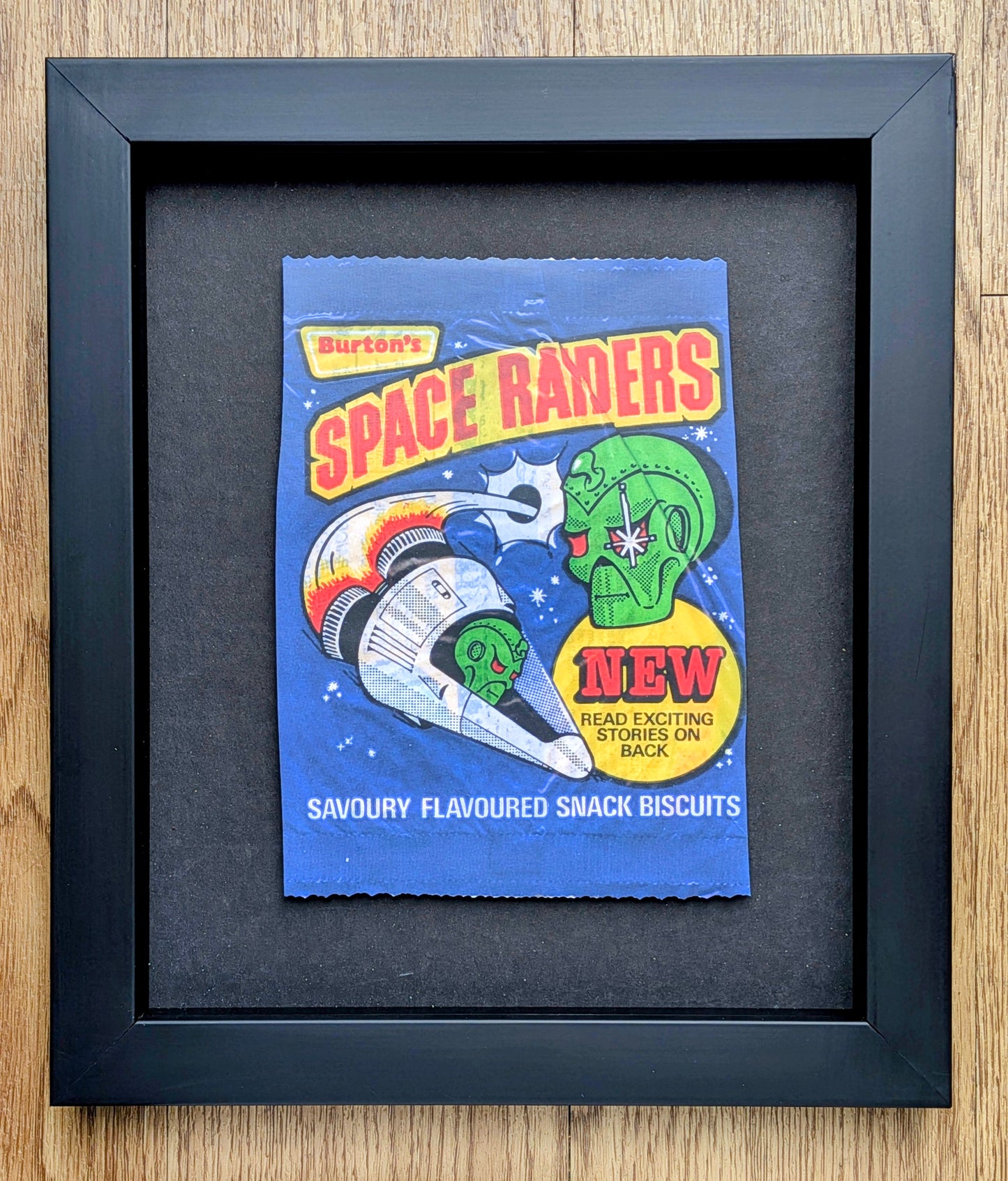Framed vintage-style advertisement for Burton's Space Raiders biscuits on a wooden surface.