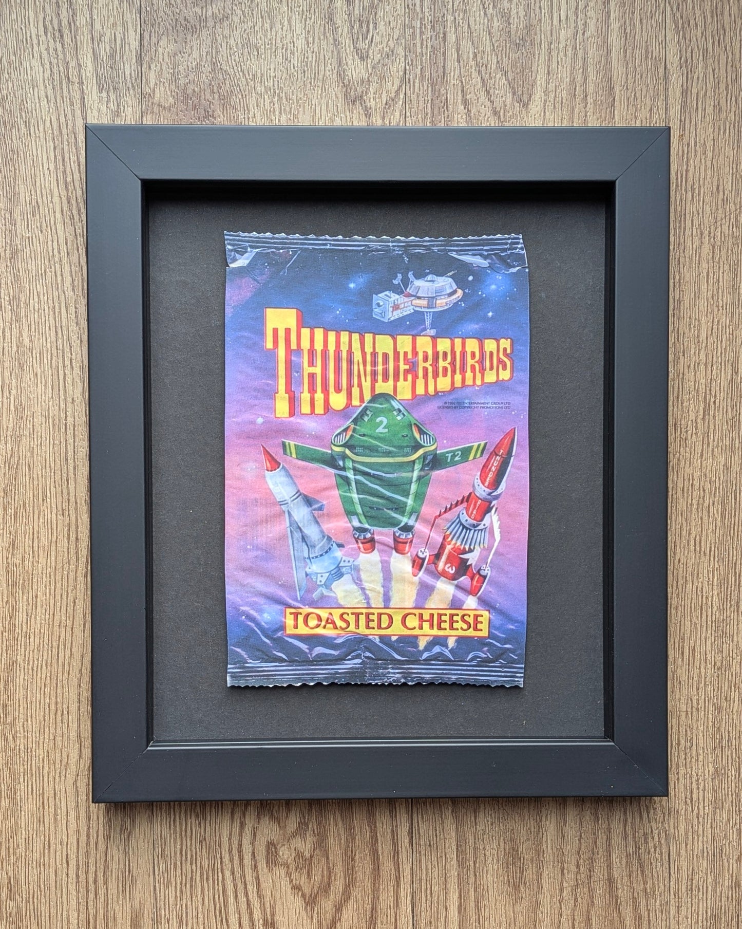 Framed vintage 'Thunderbirds' toasted cheese packaging on a wooden surface