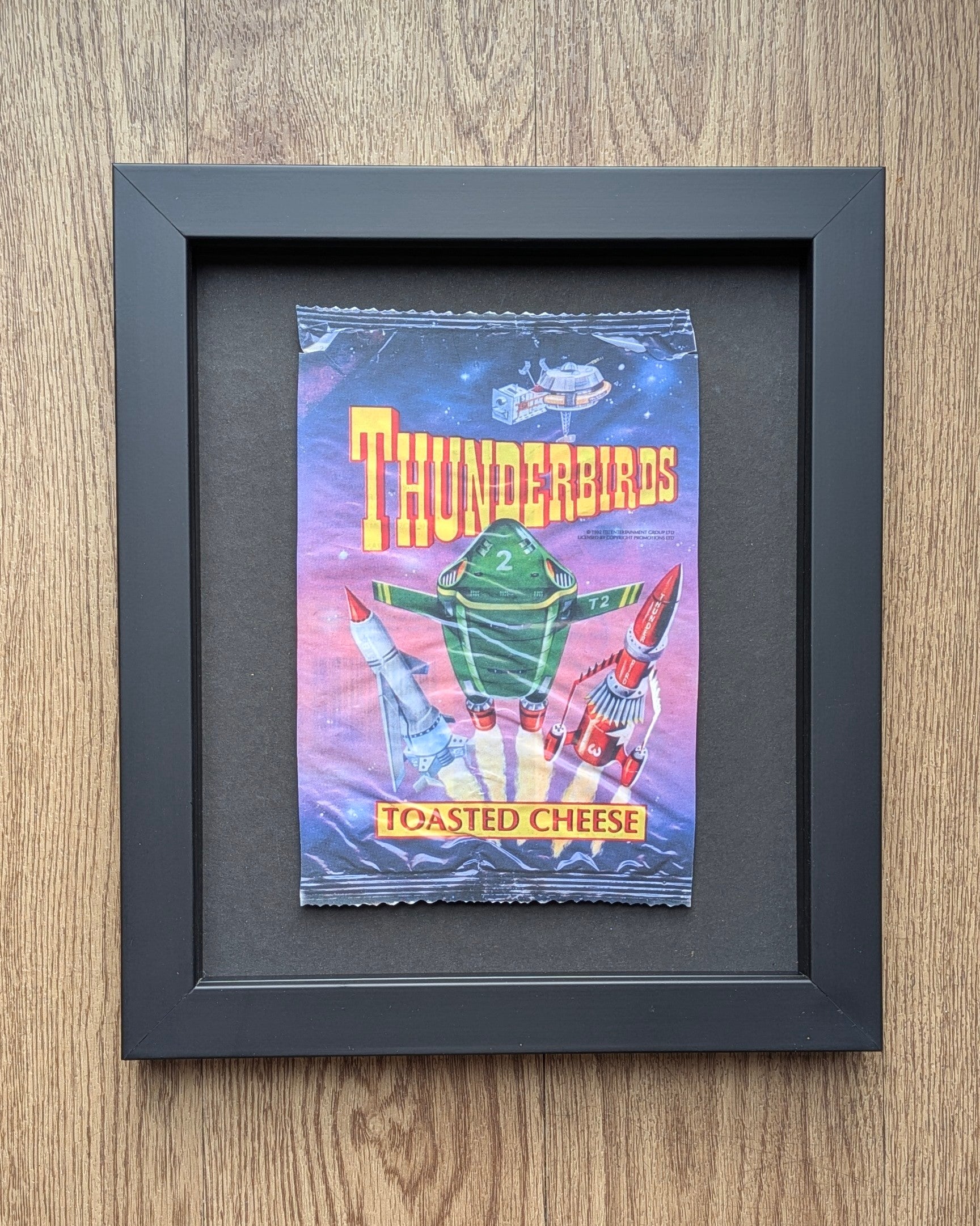 Framed vintage 'Thunderbirds' toasted cheese packaging on a wooden surface