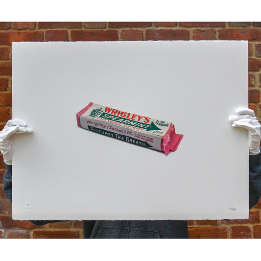 A limited edition print titled 'WRIGLEY'S SPEARMINT' by Trash Prints, featuring an artistic representation of an old gum package, being held with white gloves against brick background