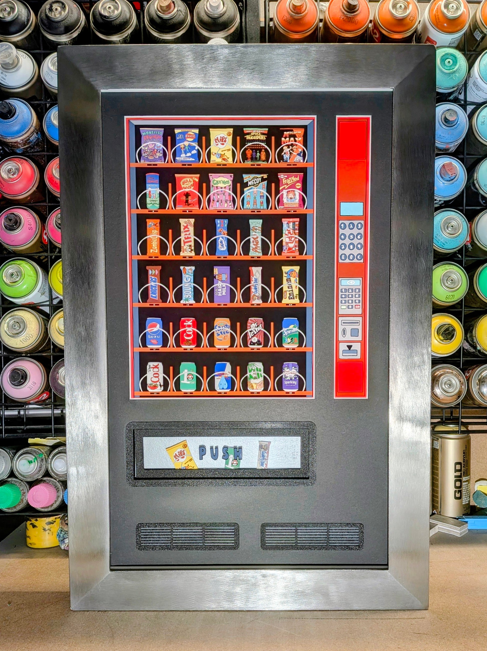 A large stainless steel vending machine picture frame with a colourful display of assorted snack and beverage packs, set against a background filled with rows of colourful spray paint cans.